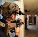 A heavily equipped tactical operator in full combat gear is conducting what appears to be a building clearance drill or training exercise in an indoor corridor. The operator is wearing a multicam helmet with night vision mount and aiming a rifle around a corner. Observers in high-visibility vests visible in the background suggest this is a supervised training or exercise scenario.