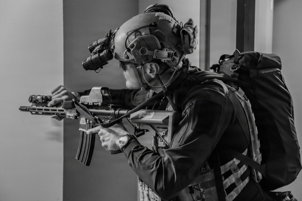 A black-and-white photograph depicts a heavily equipped tactical operator in a building environment, aiming a rifle while moving through a doorway or corridor. The individual's face has been pixelated/redacted. A second operator is partially visible in the background.