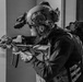 A black-and-white photograph depicts a heavily equipped tactical operator in a building environment, aiming a rifle while moving through a doorway or corridor. The individual's face has been pixelated/redacted. A second operator is partially visible in the background.