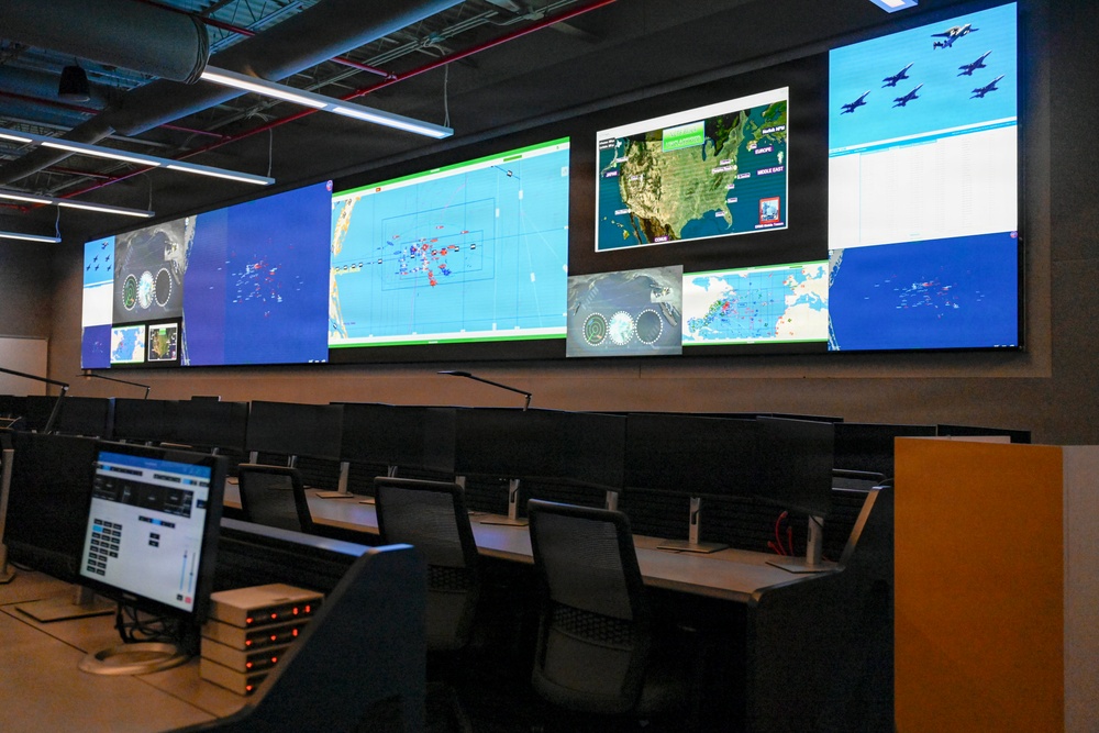 A modern operations or simulation center featuring a large multi-panel video wall displaying tactical maps, geographic overlays, and what appears to be military simulation software. The room contains rows of workstations with ergonomic chairs and individual monitors, suggesting a command, control, or training environment. The facility appears unoccupied at the time of the photograph.