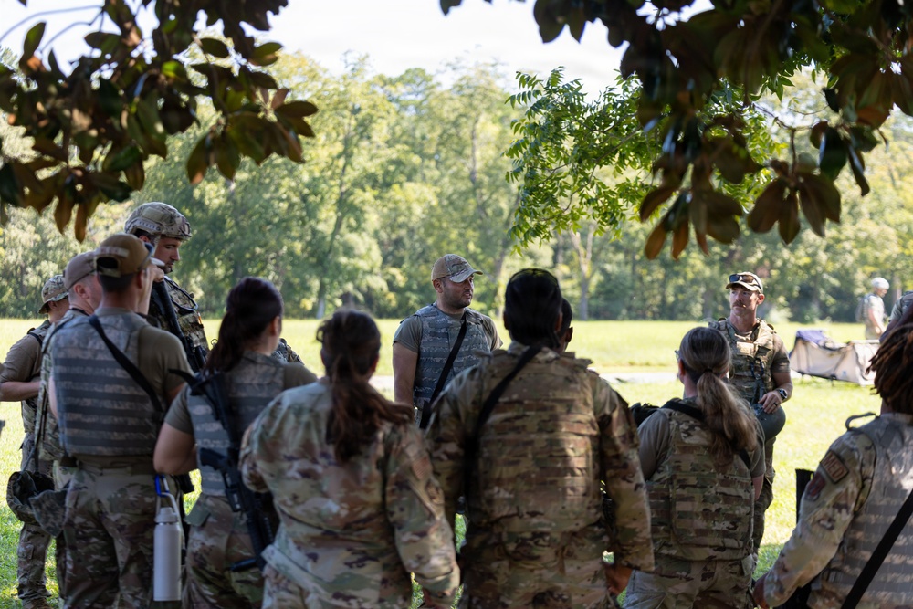A group of military personnel in camouflage uniforms are gathered in an outdoor briefing or debriefing session under tree cover in a grassy, wooded area. Two individuals appear to be addressing the group, with one wearing a tactical vest and baseball cap and another wearing full tactical gear. The setting suggests a field training exercise or operational planning session.