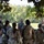 Georgia ANG Leadership Symposium 2026 A group of military personnel in camouflage uniforms are gathered in an outdoor briefing or debriefing session under tree cover in a grassy, wooded area. Two individuals appear to be addressing the group, with one wearing a tactical vest and baseball cap and another wearing full tactical gear. The setting suggests a field training exercise or operational planning session.