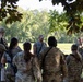 A group of military personnel in camouflage uniforms are gathered in an outdoor briefing or debriefing session under tree cover in a grassy, wooded area. Two individuals appear to be addressing the group, with one wearing a tactical vest and baseball cap and another wearing full tactical gear. The setting suggests a field training exercise or operational planning session.