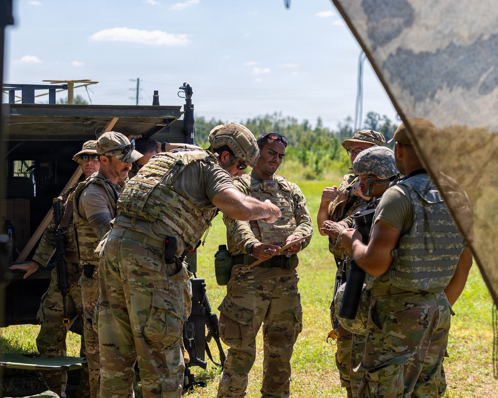 A group of military personnel in full combat gear are gathered outdoors near a military vehicle, appearing to conduct a briefing or equipment check. The soldiers are wearing multicam/OCP camouflage uniforms with plate carriers, helmets, and tactical accessories. The setting appears to be a training exercise in a grassy field with trees visible in the background.