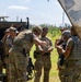 A group of military personnel in full combat gear are gathered outdoors near a military vehicle, appearing to conduct a briefing or equipment check. The soldiers are wearing multicam/OCP camouflage uniforms with plate carriers, helmets, and tactical accessories. The setting appears to be a training exercise in a grassy field with trees visible in the background.
