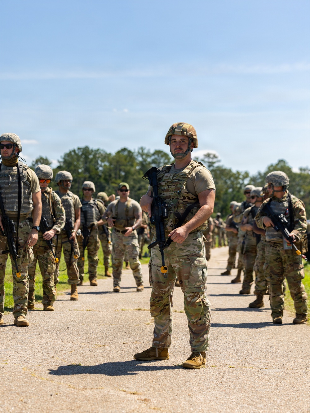 A group of military personnel in full combat gear are assembled on a paved road in an outdoor training environment. The central figure, wearing OCP (Operational Camouflage Pattern) uniform and a ballistic helmet, holds an M4-style rifle while standing at ease. The setting appears to be a military training exercise, with personnel lined up on both sides of the road.