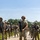 Georgia ANG Leadership Symposium 2026 A group of military personnel in full combat gear are assembled on a paved road in an outdoor training environment. The central figure, wearing OCP (Operational Camouflage Pattern) uniform and a ballistic helmet, holds an M4-style rifle while standing at ease. The setting appears to be a military training exercise, with personnel lined up on both sides of the road.