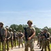 A group of military personnel in full combat gear are assembled on a paved road in an outdoor training environment. The central figure, wearing OCP (Operational Camouflage Pattern) uniform and a ballistic helmet, holds an M4-style rifle while standing at ease. The setting appears to be a military training exercise, with personnel lined up on both sides of the road.