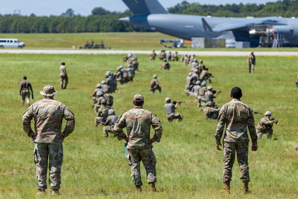 A military training exercise is taking place on an airfield grass area, with a large group of soldiers in tactical positions on the ground while three standing personnel — likely instructors or supervisors — observe from the foreground. A large military transport aircraft, consistent with a C-17 Globemaster or similar heavy-lift aircraft, is visible on the tarmac in the background. The scene suggests an airfield seizure or combat deployment drill.