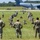 Georgia ANG Leadership Symposium 2026 A military training exercise is taking place on an airfield grass area, with a large group of soldiers in tactical positions on the ground while three standing personnel — likely instructors or supervisors — observe from the foreground. A large military transport aircraft, consistent with a C-17 Globemaster or similar heavy-lift aircraft, is visible on the tarmac in the background. The scene suggests an airfield seizure or combat deployment drill.