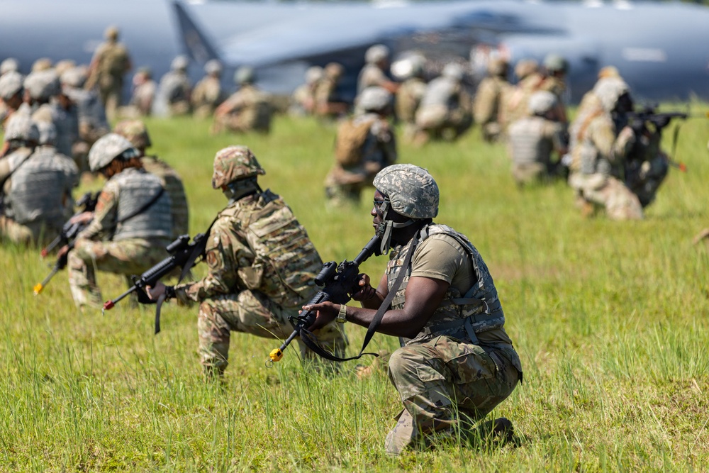 The image depicts a large-scale military training exercise taking place on an open grassy field. Multiple soldiers in camouflage uniforms and helmets are positioned in tactical stances, holding rifles with training (MILES/blank-fire) attachments. A large military aircraft is visible in the background, suggesting an airfield or landing zone scenario.