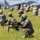 Georgia ANG Leadership Symposium 2026 The image depicts a large-scale military training exercise taking place on an open grassy field. Multiple soldiers in camouflage uniforms and helmets are positioned in tactical stances, holding rifles with training (MILES/blank-fire) attachments. A large military aircraft is visible in the background, suggesting an airfield or landing zone scenario.