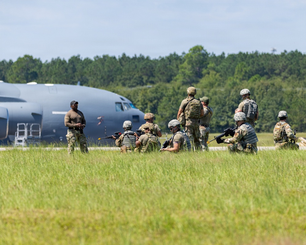 A group of military personnel in camouflage uniforms are positioned on or near an airfield tarmac, some kneeling with weapons while others stand observing. A large military transport aircraft, consistent with a C-130 Hercules, is visible in the background. The scene appears to depict a tactical training exercise or airfield security drill.