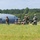 Georgia ANG Leadership Symposium 2026 A group of military personnel in camouflage uniforms are positioned on or near an airfield tarmac, some kneeling with weapons while others stand observing. A large military transport aircraft, consistent with a C-130 Hercules, is visible in the background. The scene appears to depict a tactical training exercise or airfield security drill.