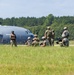 A group of military personnel in camouflage uniforms are positioned on or near an airfield tarmac, some kneeling with weapons while others stand observing. A large military transport aircraft, consistent with a C-130 Hercules, is visible in the background. The scene appears to depict a tactical training exercise or airfield security drill.