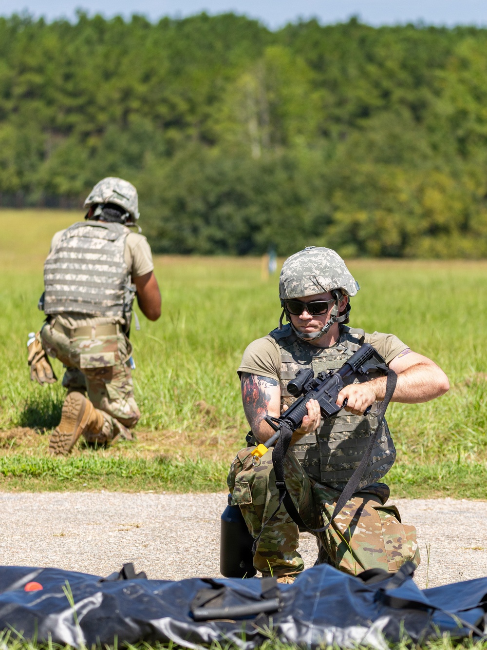 Two military personnel in full combat gear are conducting what appears to be a field training exercise on an open grassy area bordered by dense tree line. The foreground soldier is kneeling and handling an assault rifle, while the background soldier moves forward in a tactical crouch. Dark equipment or gear is visible on the ground in the foreground.