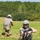 Georgia ANG Leadership Symposium 2026 Two military personnel in full combat gear are conducting what appears to be a field training exercise on an open grassy area bordered by dense tree line. The foreground soldier is kneeling and handling an assault rifle, while the background soldier moves forward in a tactical crouch. Dark equipment or gear is visible on the ground in the foreground.