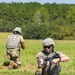 Two military personnel in full combat gear are conducting what appears to be a field training exercise on an open grassy area bordered by dense tree line. The foreground soldier is kneeling and handling an assault rifle, while the background soldier moves forward in a tactical crouch. Dark equipment or gear is visible on the ground in the foreground.