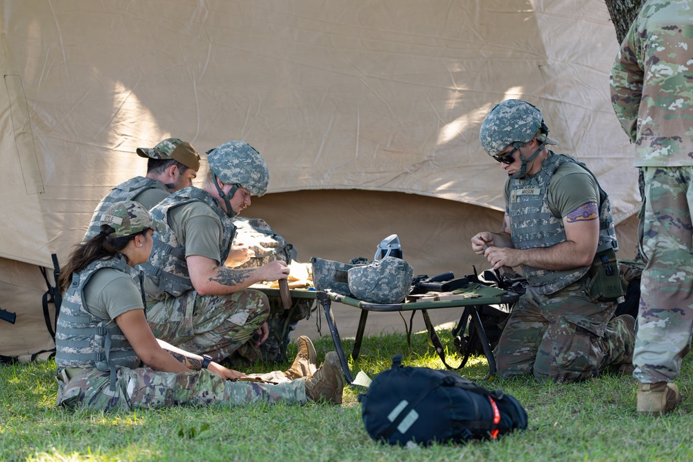 A group of military personnel in camouflage uniforms are gathered on grass near a large tan military tent, appearing to conduct a field training exercise. Several soldiers wearing body armor and helmets are clustered around a military stretcher or cot, handling equipment and gear. A fifth uniformed individual stands partially visible at the right edge of the frame.