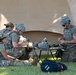 A group of military personnel in camouflage uniforms are gathered on grass near a large tan military tent, appearing to conduct a field training exercise. Several soldiers wearing body armor and helmets are clustered around a military stretcher or cot, handling equipment and gear. A fifth uniformed individual stands partially visible at the right edge of the frame.