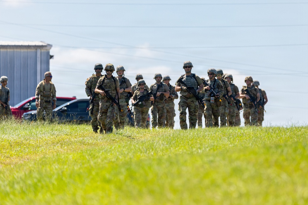 A group of approximately 15-20 armed military or tactical personnel in camouflage uniforms are moving across a grassy field. They are equipped with rifles, body armor, helmets, and tactical gear. The setting appears to be an outdoor training area or operational staging ground near a metal building structure.