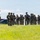 Georgia ANG Leadership Symposium 2026 A group of approximately 15-20 armed military or tactical personnel in camouflage uniforms are moving across a grassy field. They are equipped with rifles, body armor, helmets, and tactical gear. The setting appears to be an outdoor training area or operational staging ground near a metal building structure.