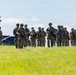 A group of approximately 15-20 armed military or tactical personnel in camouflage uniforms are moving across a grassy field. They are equipped with rifles, body armor, helmets, and tactical gear. The setting appears to be an outdoor training area or operational staging ground near a metal building structure.