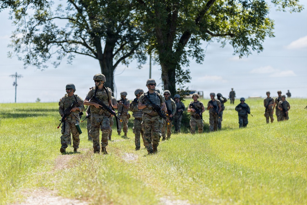 A column of military personnel in full combat gear marches along a dirt path through an open grassy field. The soldiers are equipped with rifles, tactical vests, helmets, and backpacks, suggesting a field training exercise or patrol movement. The formation stretches back into the distance with approximately 15-20 visible personnel.