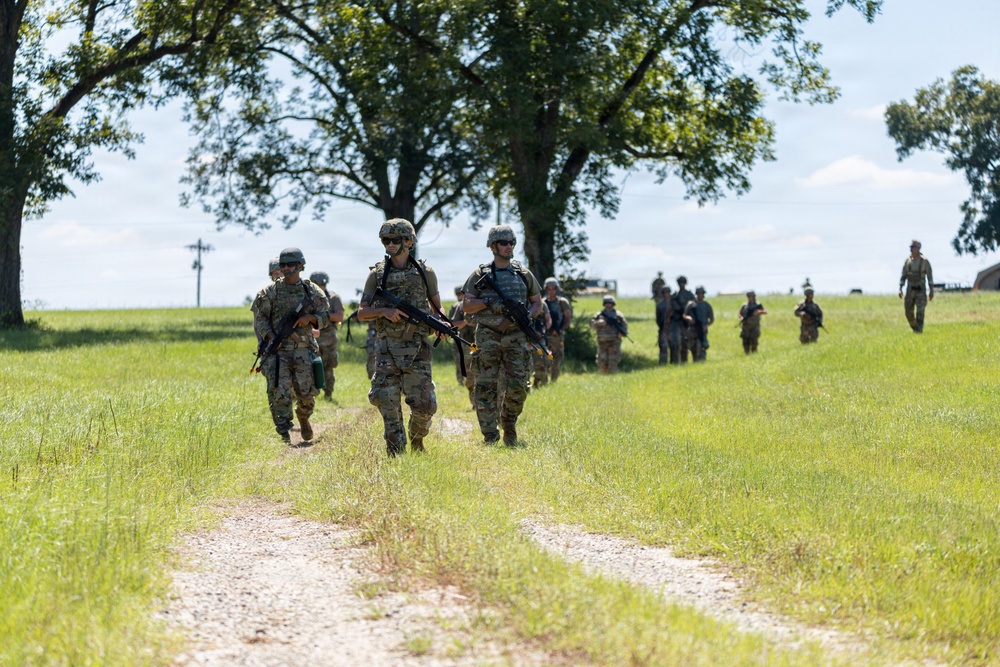 A column of military personnel in camouflage uniforms marches along a dirt path through an open grassy field. The soldiers are carrying rifles and wearing full combat gear including helmets, body armor, and backpacks. The scene appears to be a training exercise or field movement conducted in a rural outdoor environment.