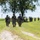 Georgia ANG Leadership Symposium 2026 A column of military personnel in camouflage uniforms marches along a dirt path through an open grassy field. The soldiers are carrying rifles and wearing full combat gear including helmets, body armor, and backpacks. The scene appears to be a training exercise or field movement conducted in a rural outdoor environment.