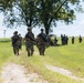 A column of military personnel in camouflage uniforms marches along a dirt path through an open grassy field. The soldiers are carrying rifles and wearing full combat gear including helmets, body armor, and backpacks. The scene appears to be a training exercise or field movement conducted in a rural outdoor environment.