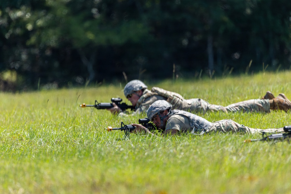 Two soldiers in military camouflage uniforms are lying prone in a grassy field in a firing position, aiming rifles toward an unseen target. Both are wearing helmets and tactical gear, suggesting a training exercise or field operation. The background consists of a tree line with dense green foliage.