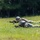 Georgia ANG Leadership Symposium 2026 Two soldiers in military camouflage uniforms are lying prone in a grassy field in a firing position, aiming rifles toward an unseen target. Both are wearing helmets and tactical gear, suggesting a training exercise or field operation. The background consists of a tree line with dense green foliage.