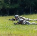 Two soldiers in military camouflage uniforms are lying prone in a grassy field in a firing position, aiming rifles toward an unseen target. Both are wearing helmets and tactical gear, suggesting a training exercise or field operation. The background consists of a tree line with dense green foliage.