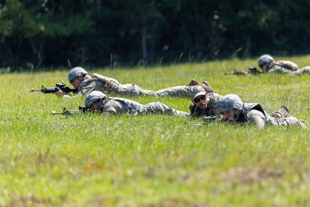 Multiple military personnel in camouflage uniforms and helmets are lying prone in a grassy field in a tactical formation. They appear to be conducting a field training exercise, with at least two individuals visibly holding rifles in the low-crawl position. The background consists of dense green treeline under bright daylight conditions.