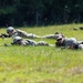 Multiple military personnel in camouflage uniforms and helmets are lying prone in a grassy field in a tactical formation. They appear to be conducting a field training exercise, with at least two individuals visibly holding rifles in the low-crawl position. The background consists of dense green treeline under bright daylight conditions.