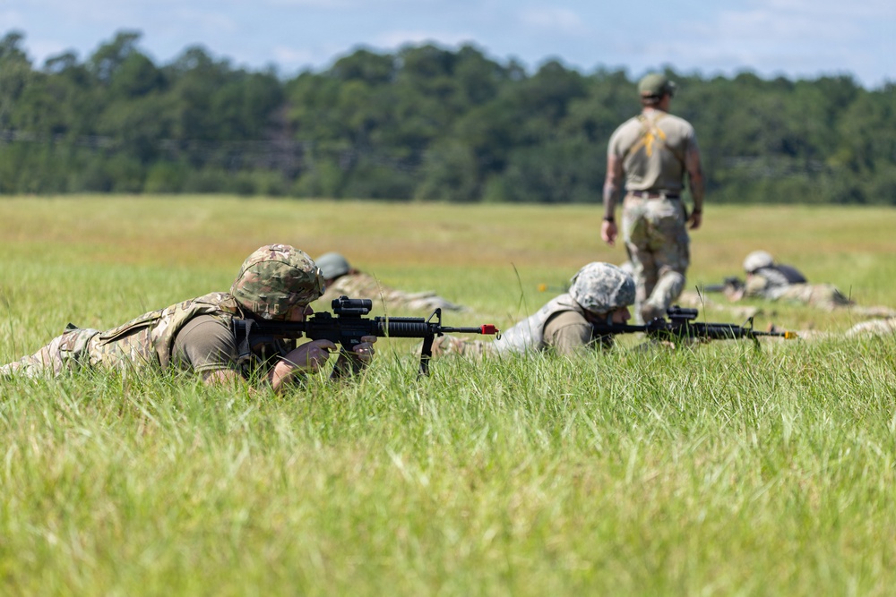 Military personnel are conducting a prone firing exercise in an open grassy field. Multiple soldiers are lying in the low crawl position aiming rifles downrange while a standing instructor or cadre member observes from behind. The setting appears to be a training range, likely at a military installation.