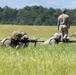 Military personnel are conducting a prone firing exercise in an open grassy field. Multiple soldiers are lying in the low crawl position aiming rifles downrange while a standing instructor or cadre member observes from behind. The setting appears to be a training range, likely at a military installation.