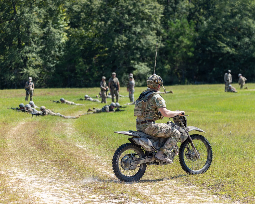 A military training exercise is taking place in an open grassy field bordered by dense trees. A soldier in full camouflage gear and helmet is mounted on a dirt motorcycle on a dirt track, observing the exercise. Multiple soldiers are visible in various tactical positions including prone firing positions and standing formations across the field.