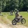 Georgia ANG Leadership Symposium 2026 A military training exercise is taking place in an open grassy field bordered by dense trees. A soldier in full camouflage gear and helmet is mounted on a dirt motorcycle on a dirt track, observing the exercise. Multiple soldiers are visible in various tactical positions including prone firing positions and standing formations across the field.