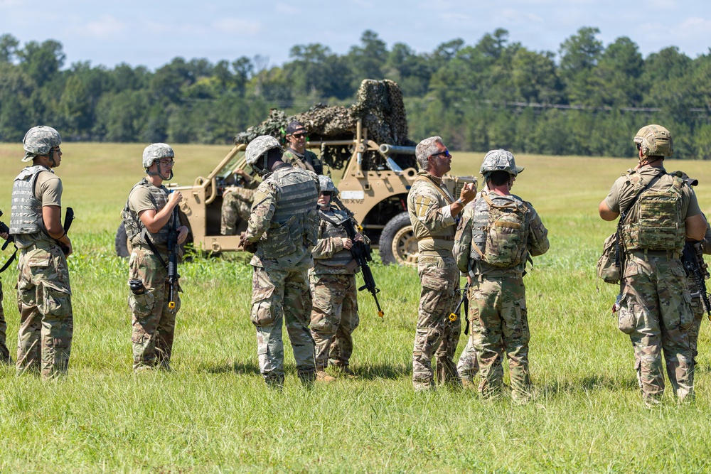 A group of approximately seven military personnel in camouflage uniforms are gathered in an open grassy field, appearing to conduct a briefing or debrief. A tan military tactical vehicle covered with camouflage netting is visible in the background. The setting suggests a training exercise or field operation.