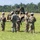 Georgia ANG Leadership Symposium 2026 A group of approximately seven military personnel in camouflage uniforms are gathered in an open grassy field, appearing to conduct a briefing or debrief. A tan military tactical vehicle covered with camouflage netting is visible in the background. The setting suggests a training exercise or field operation.