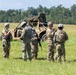 A group of approximately seven military personnel in camouflage uniforms are gathered in an open grassy field, appearing to conduct a briefing or debrief. A tan military tactical vehicle covered with camouflage netting is visible in the background. The setting suggests a training exercise or field operation.