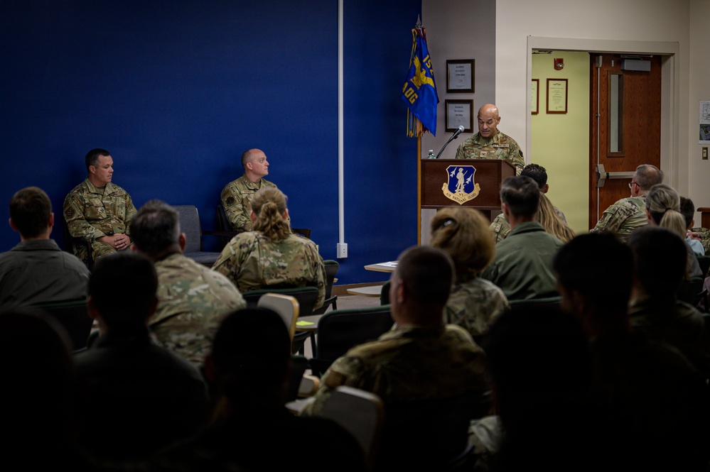 A military briefing or ceremony is taking place in what appears to be a conference or meeting room at an Air Force installation. A uniformed service member stands at a wooden podium bearing an Air Force unit emblem, addressing an audience of seated military personnel in camouflage uniforms. Two additional uniformed individuals are seated on the stage area to the left of the speaker.