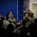 A military briefing or ceremony is taking place in what appears to be a conference or meeting room at an Air Force installation. A uniformed service member stands at a wooden podium bearing an Air Force unit emblem, addressing an audience of seated military personnel in camouflage uniforms. Two additional uniformed individuals are seated on the stage area to the left of the speaker.