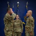 Three U.S. Air Force personnel in OCP (Operational Camouflage Pattern) uniforms are participating in a formal military ceremony, likely a change of command or assumption of command. Two personnel on the left and right are exchanging salutes while a third stands at attention in the center holding a guidon staff. The setting features a blue wall backdrop with the U.S. flag, Air Force flag, and what appears to be a unit flag.