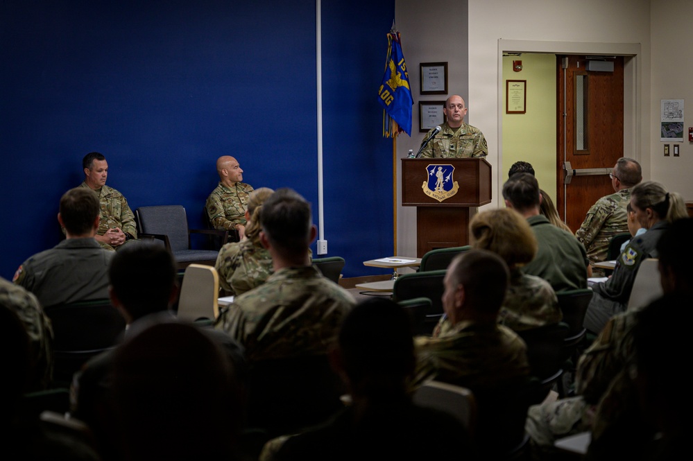 A military briefing or ceremony is taking place in a conference room, with a uniformed officer speaking at a podium bearing a military unit insignia. An audience of military personnel in camouflage uniforms is seated facing the speaker, while two additional officers are seated to the left of the podium. A blue unit guidon flag is visible in the background.