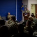 A military briefing or ceremony is taking place in a conference room, with a uniformed officer speaking at a podium bearing a military unit insignia. An audience of military personnel in camouflage uniforms is seated facing the speaker, while two additional officers are seated to the left of the podium. A blue unit guidon flag is visible in the background.