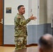 A U.S. Army soldier in OCP (Operational Camouflage Pattern) uniform is presenting or briefing to an audience in what appears to be a military training or conference room. He is gesturing with his right hand while speaking, with a readiness-themed banner visible in the background. The banner covers topics including Physical Readiness, Mental Readiness, Spiritual Readiness, Nutritional Readiness, and Sleep Readiness.
