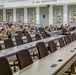 A military lecture hall or auditorium labeled 'THE INSTITUTION' is filled with a mix of uniformed military personnel and civilians attending a briefing or presentation. The tiered seating arrangement and formal setup suggest an educational or training environment, likely at a U.S. Army school or institution. Several informational banners and unit insignia displays line the walls, reinforcing the military educational context.
